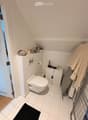 Compact bathroom with white tiled floor and walls, toilet, small cabinet, mirror, towel, and personal care items on shelves.