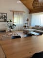 Modern living room with wooden floors, a stone fireplace, large windows with white curtains, a grey sectional sofa, a glass coffee table, a wooden dining table with black chairs, a round ottoman, a cat tree, a floor lamp, and decorative shelves with plant