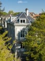 A multi-story house with a grey roof, white walls, and ornate architectural details, surrounded by trees and greenery.