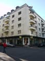 Multi-story residential building with balconies and storefronts at ground level, located on a street with parked cars and pedestrians.