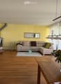 Bright living room with wooden flooring, a beige sectional sofa, a wicker armchair, a glass coffee table, a large flower vase on a wooden side table, and a modern floor lamp.