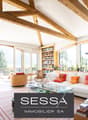 Bright living room with exposed wooden beams, large windows offering a scenic view, a white sectional sofa with colorful cushions, a glass coffee table, a wooden chair, a bookcase, and decorative items including a candle holder and a statue.