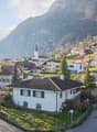 A picturesque village scene with traditional houses, a church tower, and surrounding mountains under a clear blue sky.