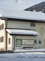 A two-story building with snow-covered roof and red shutters, located in a snowy village with mountains in the background.