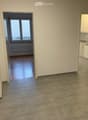Empty hallway with tiled floor and wooden flooring in adjacent rooms, featuring white walls and large windows.