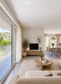 Modern, open-concept living and dining area with large windows offering a scenic view, featuring a beige sofa, wooden coffee table, flat-screen TV, dining table with chairs, and a sleek kitchen island.