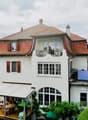 A multi-story white house with red-tiled roof, multiple windows, and a balcony. Solar panels are installed on the roof, and there is a covered outdoor seating area with tables and chairs.