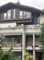 Modern two-story house with large windows, balconies, and surrounding greenery.