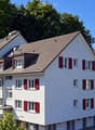 A modern, multi-story residential building with white walls, red shutters, and a tiled roof, surrounded by greenery and trees.