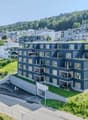 modern apartment building, blue facade, multiple balconies, green roof, raised ground floor, underground parking area, located on a hill, surrounded by greenery