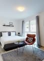 Bedroom with bed, pillow, blanket, side tables with lamps, chair, table with chairs and plates, carpet, white walls, windows, gray curtains, framed pictures, ceiling lamp, kitchen with oven, coffee machine, plate, glasses