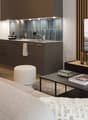 The image shows an open-concept living room and kitchen area. The kitchen features dark cabinets, a tile backsplash, and stainless steel appliances. The living room has a large sofa, a coffee table, and a TV mounted on the wall. The room also has a large