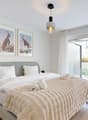 modern bedroom with gray headboard, two single beds with white linens, two white pillows, gray blanket on bed, two lamps, framed pictures of animals, ceiling light, white walls, floor to ceiling windows, balcony with white curtains, terrace with greenery