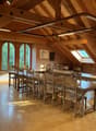 Large open-concept dining room with wooden beamed ceiling, arched windows overlooking the surrounding forest, wooden tables and chairs, and hardwood flooring.