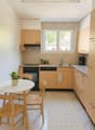 A modern and minimalistic kitchen with white walls and tiled flooring. The kitchen features a white round table and chairs, a rug, wooden cabinets, an oven, and a coffee machine. There is a window with a view outside.