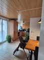 Dining area with wooden table, chairs, and a high chair. Wooden cabinets, kitchen appliances, and a window with curtains. Potted plants on the floor.
