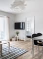 Living room with white walls, wooden floor, glass door to balcony, TV mounted on wall, dining table with 4 chairs, small side table with plant, white rug, lamp, picture frame on wall