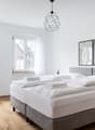Spacious bedroom with white walls, large windows, hardwood floors, a gray upholstered bed frame, white bedding, and a geometric pendant light fixture. The room also features potted plants and framed artwork on the walls.