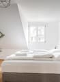 Spacious bedroom with white walls, hardwood floors, large windows, a king-size bed with white bedding, a potted palm plant, and a modern geometric light fixture. The room also features a small side table and a framed mirror on the wall.