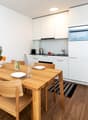 A wooden dining table with 4 chairs, white kitchen with oven, fridge, and sink