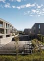 Multi-story apartment building with modern, minimalist design featuring glass windows and balconies. The building is surrounded by greenery and landscaping.
