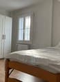 The image shows a bedroom with a double bed, a white wardrobe, and a wooden chair. The room has a window that provides natural light, and the walls are painted in a light, neutral color.