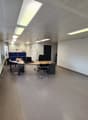 The image shows a modern, open-plan office space with a tiled floor, suspended ceiling with recessed lighting, and several desks with computers and office chairs. The space appears to be well-lit and organized, with a Proman sign visible in the corner.