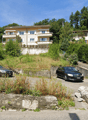 2-story apartment building with balconies, surrounded by trees and greenery, with a parking area in the foreground containing two cars