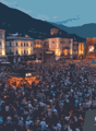 A large, crowded town square at night with brightly lit buildings and a large gathering of people