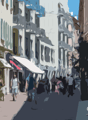 Busy urban street with shops, pedestrians, tall buildings, trees, awnings, and shadows