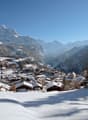 Mountain village with wooden houses, snow-covered roofs, surrounded by snowy mountains, pine trees, and a clear blue sky.