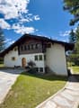 2-story chalet-style house with wooden beams, white walls, and large windows. The house is surrounded by a lush green lawn and pine trees, with a stone pathway leading to the entrance. There appears to be a balcony or terrace on the upper level.