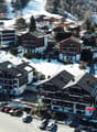 Aerial view of a ski resort town with snow-covered buildings, chalets, and a parking lot. The town is surrounded by snowy mountains and forests.