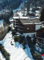 Aerial view of a snowy ski resort town with residential buildings, ski slopes, and a parking area visible.