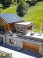 2-story wooden and stone house with a slanted roof, large balcony, garage, and parking space. The house is surrounded by a lush green landscape with trees and vegetation.