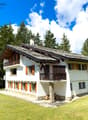 Alpine-style house, white exterior, wooden balcony, green roof, large windows, surrounded by a green lawn and trees