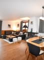 Spacious open-plan living area with hardwood floors, white walls, large windows, black leather sofa, wooden dining table and chairs, fireplace, and decorative lighting fixtures