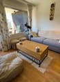Cozy living room with large windows, gray sofa, wooden coffee table, and a telescope on a tripod. The room has a balcony or terrace with a scenic mountain view.
