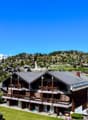 Multi-story wooden chalet-style building with balconies, surrounded by a grassy area and pine trees, with a village and mountains in the background.