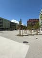 The image shows a modern residential area with multi-story apartment buildings. The buildings have balconies and are surrounded by a paved plaza with trees and benches. The area appears to be well-designed and maintained, with a clean and open layout.