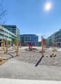 The image shows a public plaza or outdoor space in an urban setting. The area is paved with gravel and features various wooden structures, possibly seating or play equipment. The surrounding buildings have balconies and appear to be modern, multi-story re