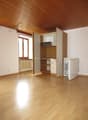 Empty room with wooden floor, white walls, kitchen area with open cabinets, window, and a refrigerator.