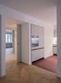 Minimalist modern kitchen, white cabinets, wooden floor, red tiles near oven, sliding door leading to another room, open window in other room.