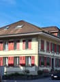 Building has brown tiled roof, yellow walls, and red shutters on windows, garden area with plants, bench, and a car parked nearby
