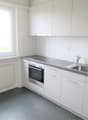 Modern kitchen with white cabinets, stainless steel countertop, built-in oven, and a sink with a faucet. It has a window with a white frame. The floor is tiled with gray tiles.