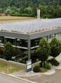Commercial building with solar panels, multiple cars visible in windows, many cars parked outside, landscape with farm and trees