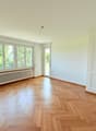 Spacious room with large windows, hardwood parquet flooring, and a radiator. The room appears to be empty, with white walls and a door leading to another area.