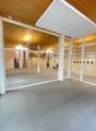 The image shows a large, open commercial space with a wooden ceiling and walls. The space has a grey carpeted floor and large glass windows that allow natural light to enter. There are various lighting fixtures and equipment visible, suggesting this could
