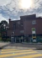A multi-story commercial building with a Migros grocery store and other businesses on the ground floor. The building has a modern, dark-colored exterior with large windows. There are trees and landscaping around the building, and a crosswalk in front of t