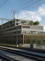 Modern office building with glass facade, located near a railway track, surrounded by trees and a house